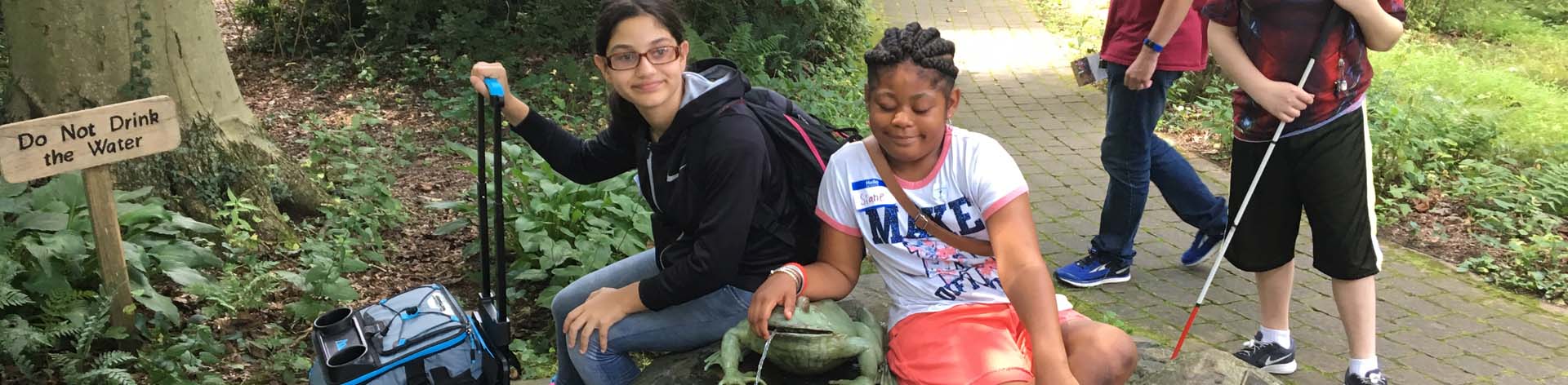 Two NFBDE BELL participants smiling together at a nature center. One girl has her arm around stone frog that is part of a fountain.