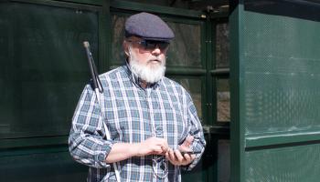 A senior man stands at a bus stop using his phone to listen to the latest news on NFB-NEWSLINE.